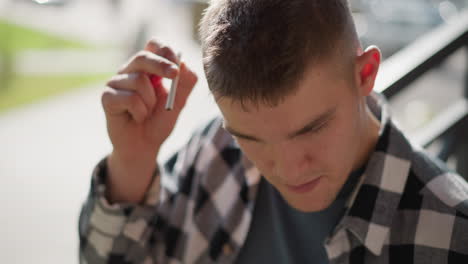 top down view of student in plaid shirt on balcony metal staircase pauses removing cigarette from ear flicks lighter ignites tip with sunlit sparks against blurred bokeh and greenery backdrop