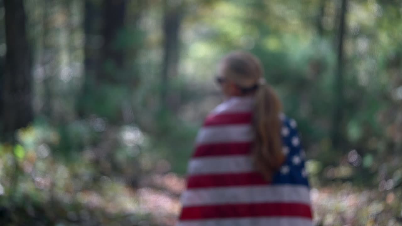 movimiento lento dentro y fuera de enfoque como mujer envuelta en una bandera americana camina por un camino en un bosque