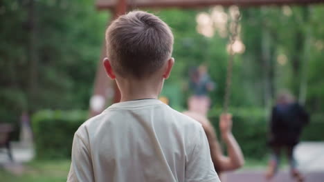back view of young boy gently pushing friend on wooden chain swing in sunlit park playground at dusk and slide in background capturing playful sibling moment and carefree summer fun