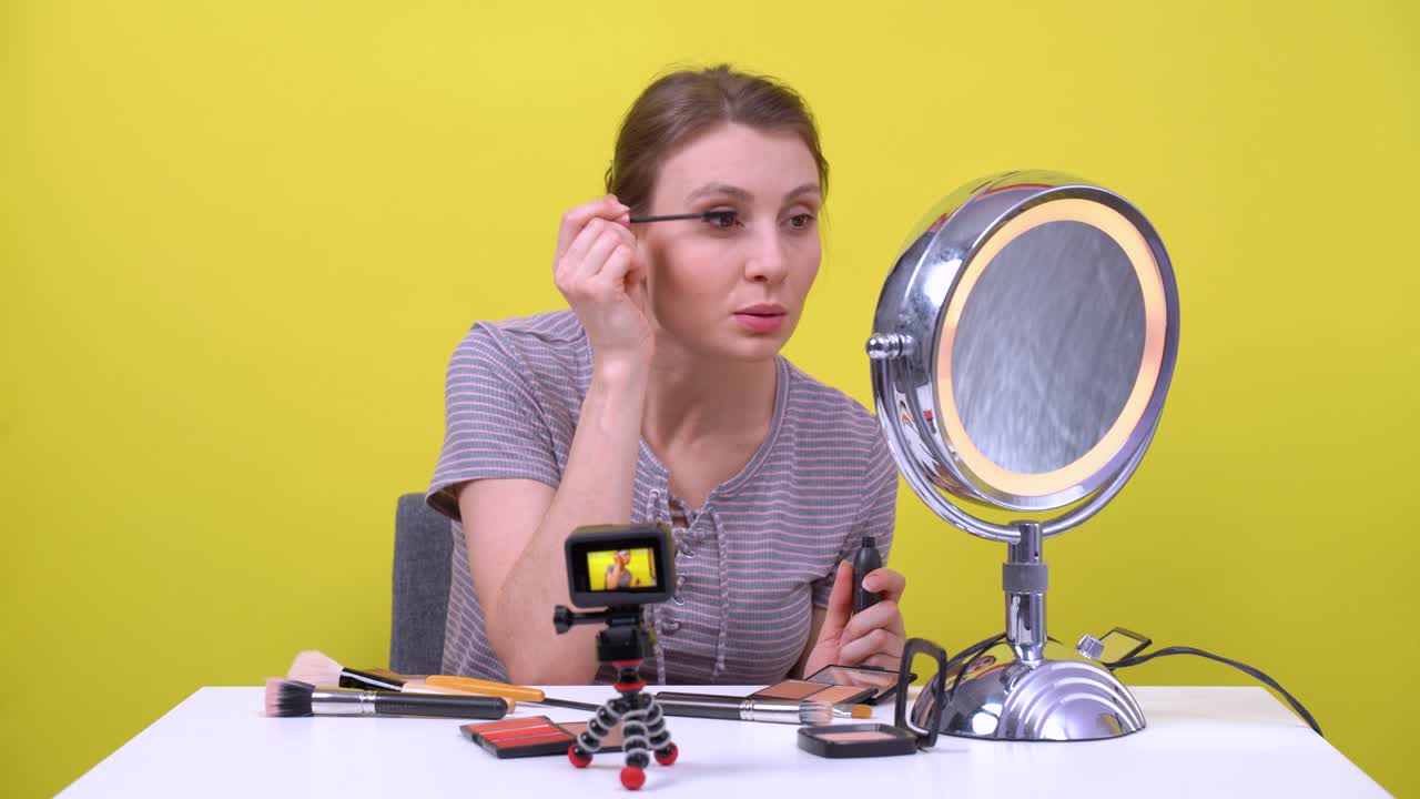 Young freelancer brown haired girl paints eyelashes with mascara in front of round mirror, talks, laughs and records beauty vlog or streams live on action camera in the studio, yellow background.