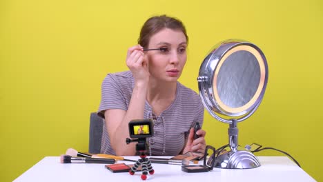young freelancer brown haired girl paints eyelashes with mascara in front of round mirror, talks, laughs and records beauty vlog or streams live on action camera in the studio, yellow background.