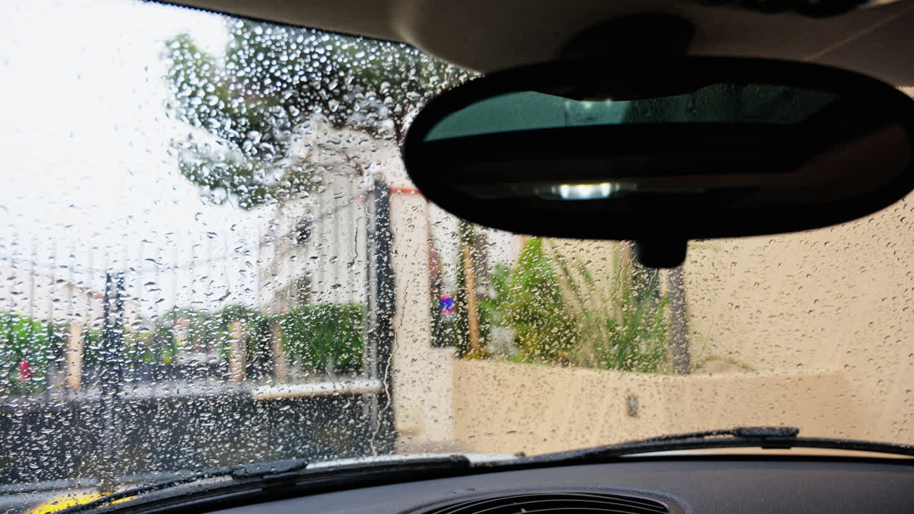 Primo piano di gocce di pioggia che cadono sul parabrezza di un'auto con vista su un cancello, una recinzione e degli alberi
