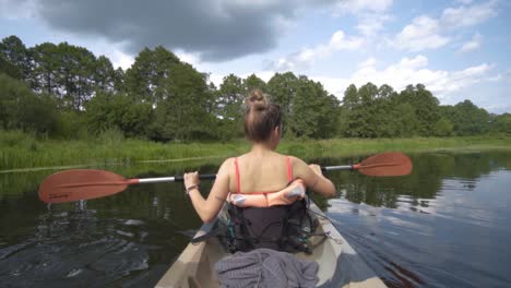 blonde girl with her back to the camera paddling a kayak in slow motion on a river surrounded by forest