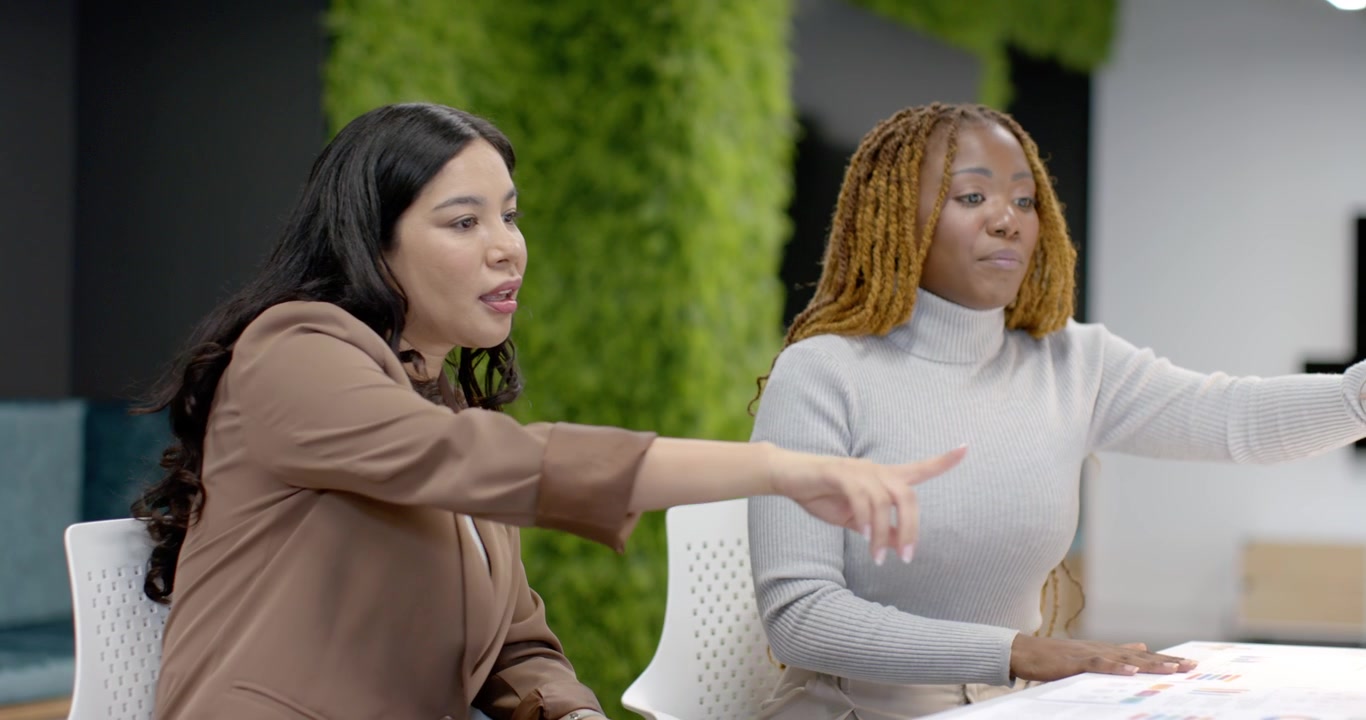 Diverse female coworkers gray-sweater placing hand on plan, pointing and reviewing at office table