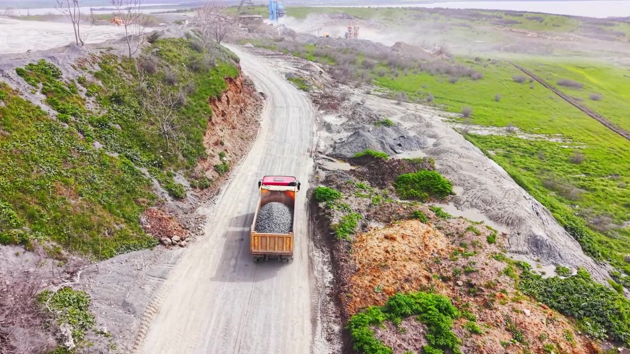 Dumper carico che guida su strada sterrata della cava. Vista aerea di carico di ghiaia, vegetazione verde del fianco della collina e impianto di lavorazione distante.