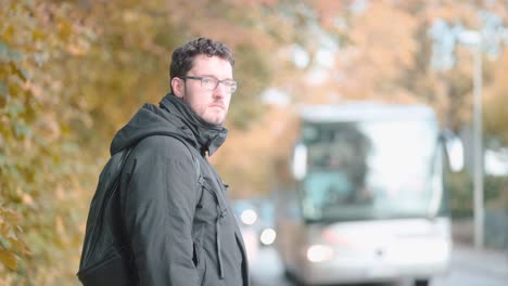 Young,-modern-German-man-with-distinctive-beard-and-glasses-calmly-stands-in-a-bustling-suburb,-observing-the-vibrant-traffic-of-buses-and-other-vehicles-as-they-pass-by