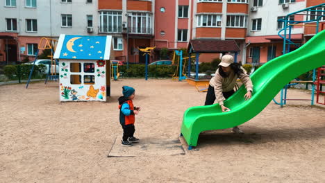 mother plays with her baby boy on the playground. woman rolls the ball by the slide and kid runs by the toy.
