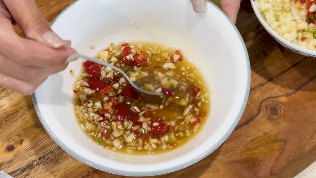 Hands use a spoon to mix chopped garlic, red chili, and fish sauce in a white bowl on a wooden table under bright, natural lighting
