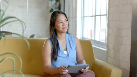 biracial woman sits comfortably on a yellow sofa, holding a tablet