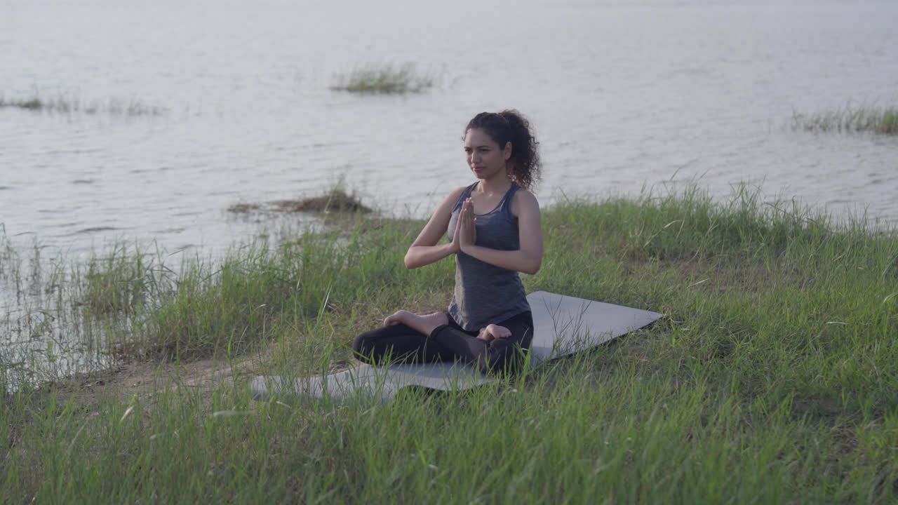 A slow-motion of a South Asian Woman in Lotus Pose (Padmasana) with prayer hands meditating by a lake on a mat