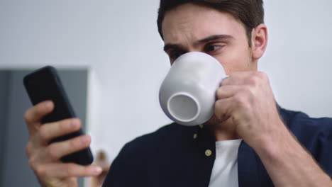 Portrait-of-young-man-typing-mobile-phone-at-home-kitchen.-Guy-drinking-coffee