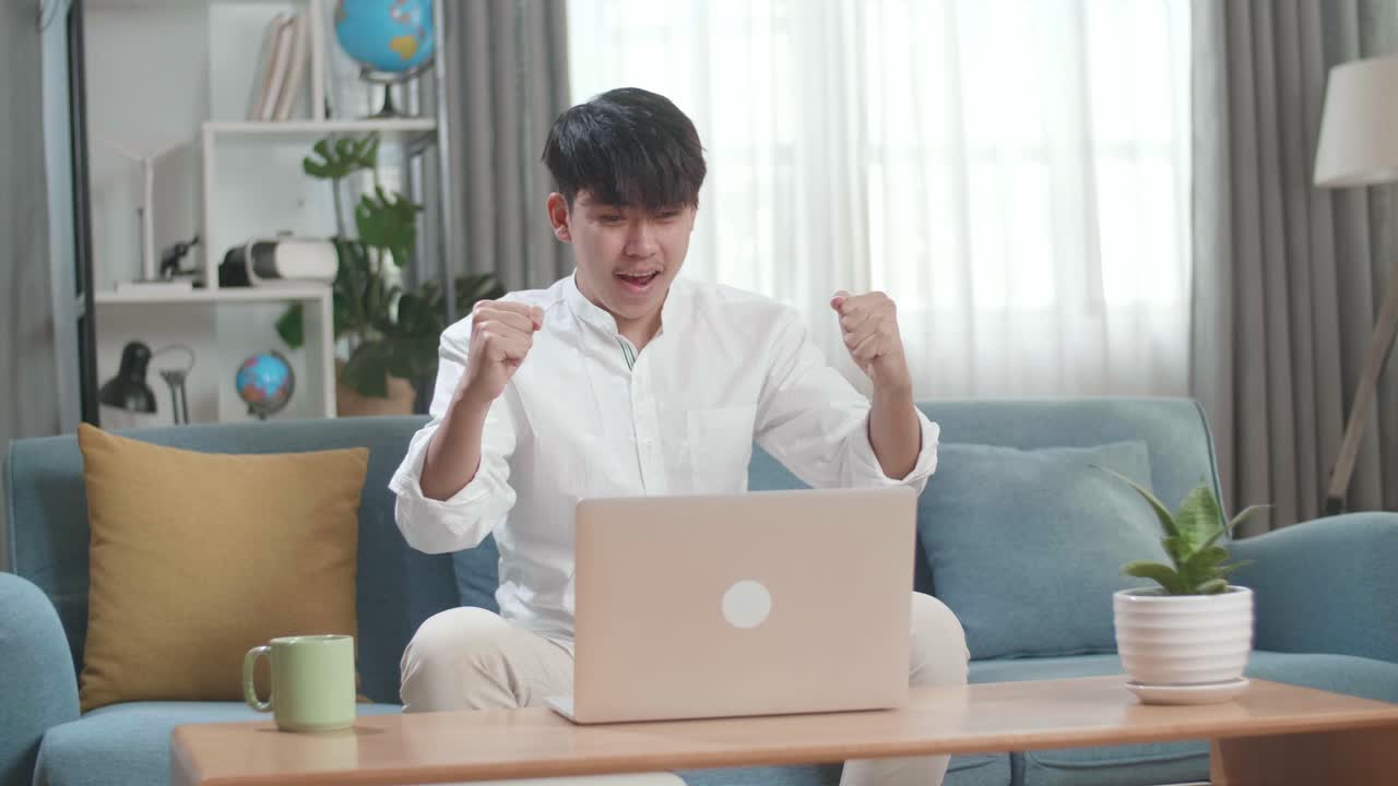 Happy Asian Man Celebrating While Using Laptop Computer In The Living Room