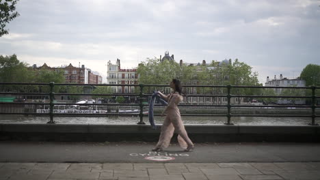 fashionable latina model with black wavy hair and a jean jacket walking in london with a view of boats and buildings behind her