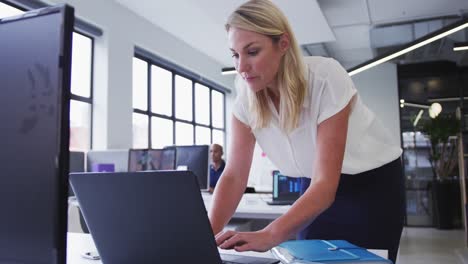 caucasian businesswoman standing using laptop in office with coworker in background
