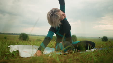 woman with grey hair lying one sided on yoga mat practicing side bend pose, arms extended, eyes closed, rise up slowly, under cloudy skies in a serene grassy field with trees in the distance