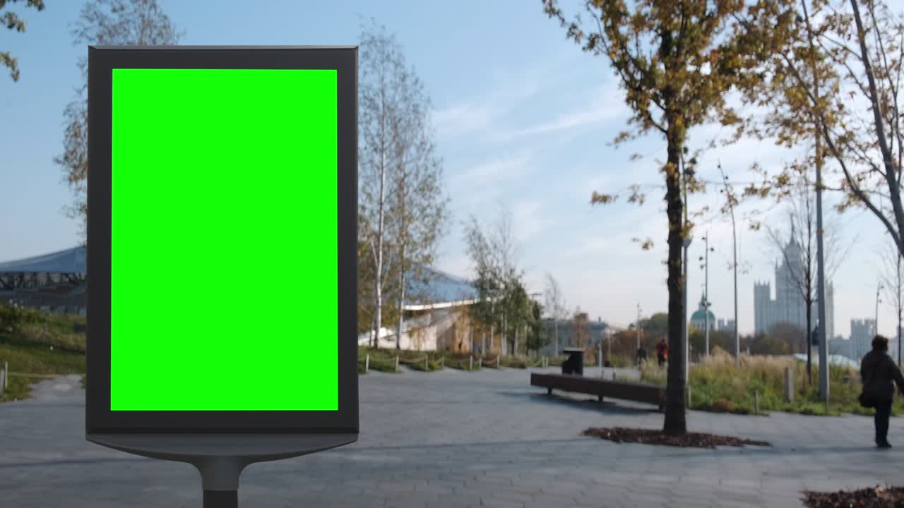 Empty billboard with a green screen for advertising in the park on the beautiful square with trees, autumn sunny shot