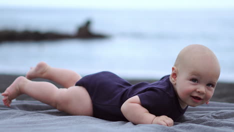 Positioned on his stomach on the black sand next to the ocean, the baby bursts into laughter while meeting the camera&#039;s gaze