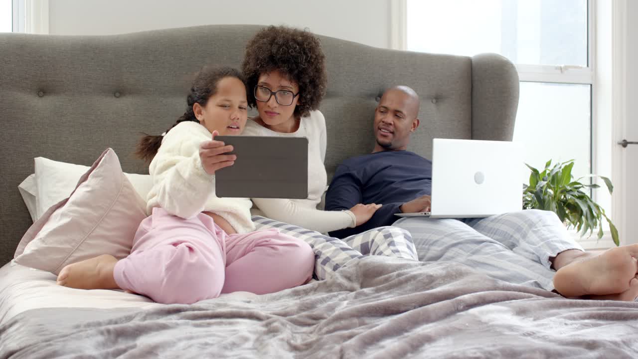Diverse family daughter frowning at tablet, lifting it to parents on bed, sparking shared scrolling