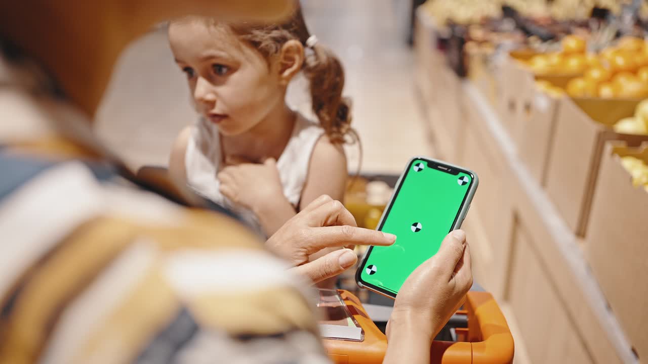 Close-up of an unrecognizable woman in a supermarket while shopping looks at the smartphone screen, while her little daughter is sitting in a supermarket cart. Chroma key on the phone screen