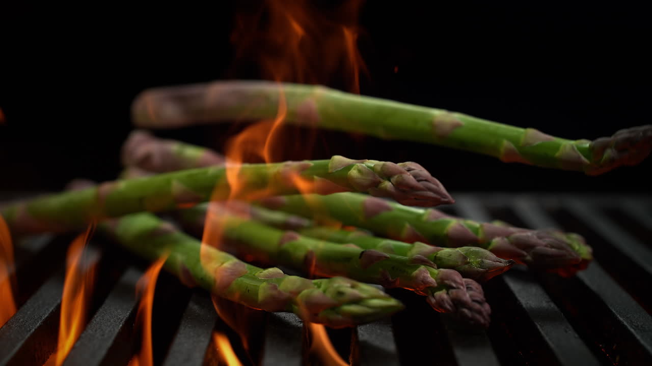 Asperges vallen op een houtskoolgrill tegen een zwarte achtergrond, vlammen barsten onder het grillrooster vandaan in slow motion