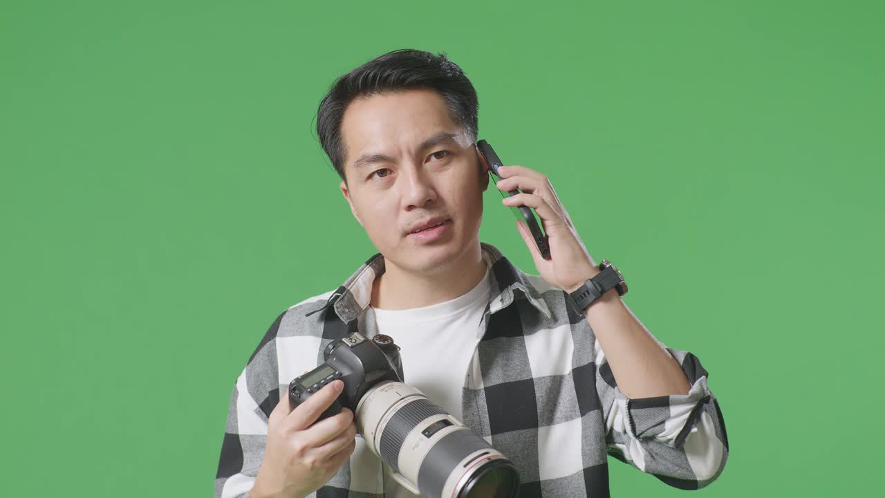 스튜디오에서 녹색 화면 배경에 서 있는 동안 손에 카메라를 들고 스마트폰으로 대화하는 아시아 사진작가의 클로즈업