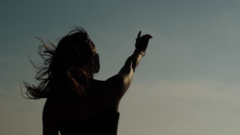 a woman raises her hand to the sky as her hair blows in the coastal wind, captured in silhouette along portugal’s shoreline
