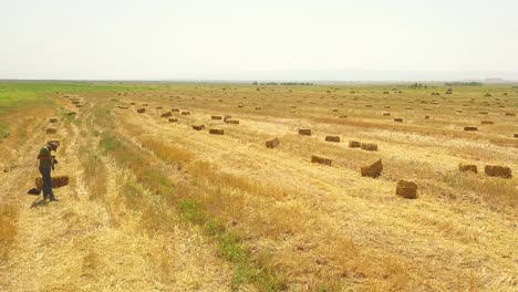 Fly-over-a-photographer-man-with-camera-and-crop-golden-wheat-or-rye-field-with-stook-hay-straw-bales