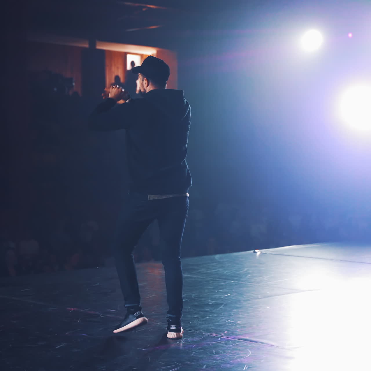 Hip-hop dancer in black cap and casual clothes dances on stage in the club. Modern dance class on scene for young audience in the studio indoors. Dancing concert.