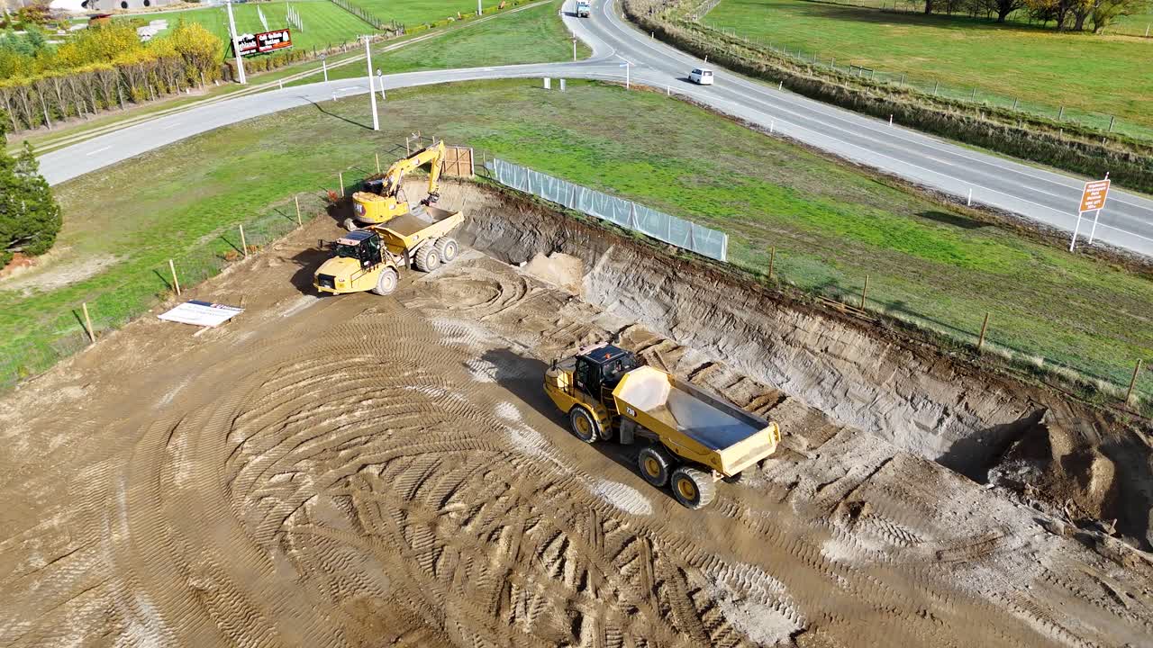 Drone footage captures an excavator and dump truck working on a construction site under bright daylight in Cromwell, New Zealand