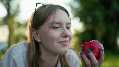 close up of lady smiling softly while taking a gentle bite from a red apple, with a beautifully blurred green background, enhancing a moment of relaxation and enjoyment in nature