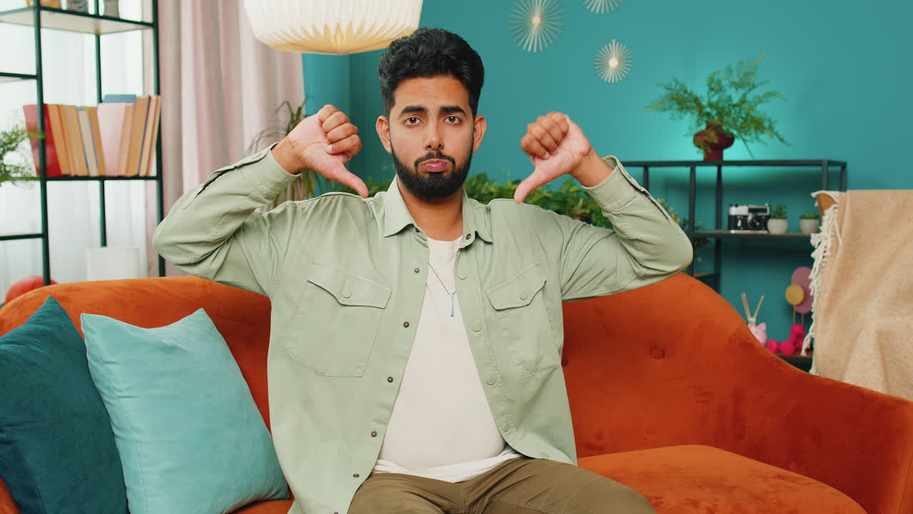 화난 아랍인 남자가 엄지손가락을 내려놓고, 나쁜 일을 싫어하고, 승인하지 않고, 집에서 불만족스러운 피드백을 합니다.