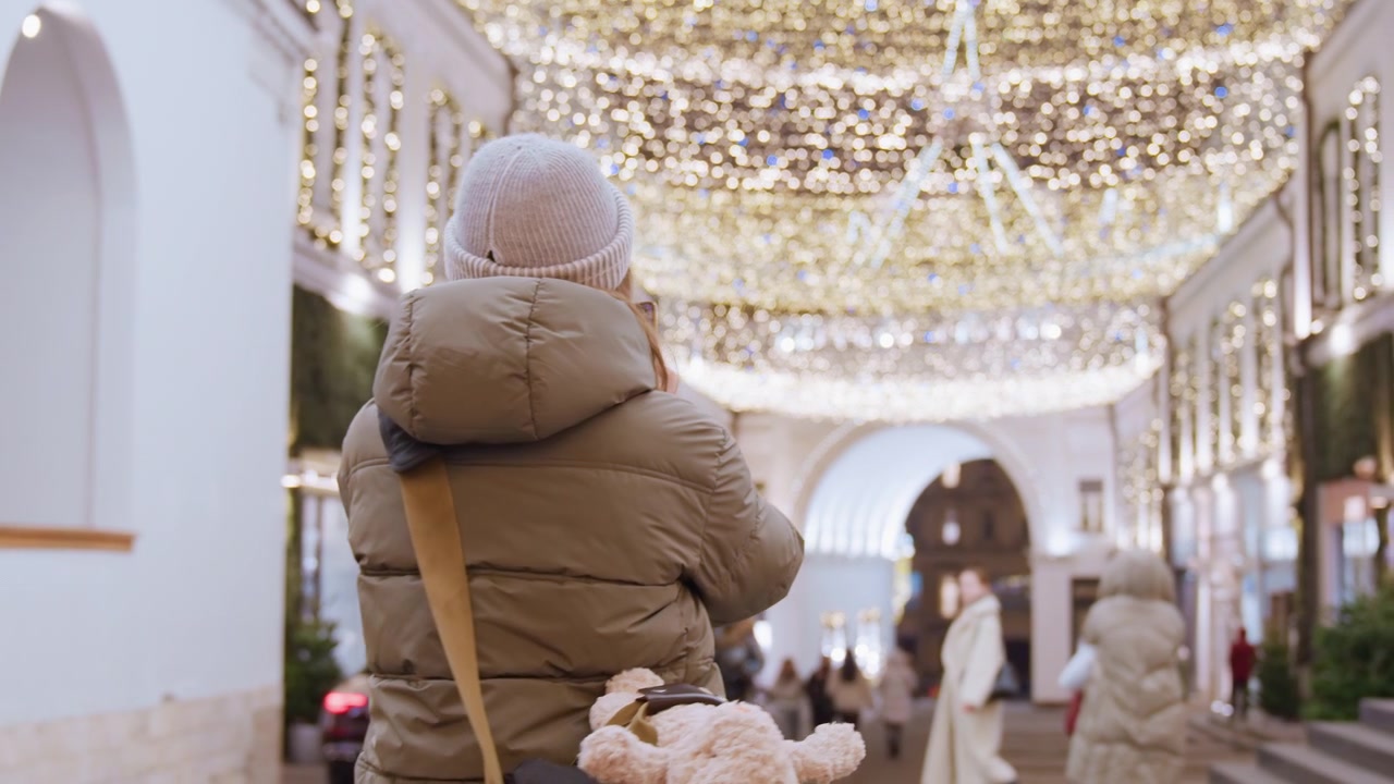 Acquirente sotto le luci festive a soffitto, piumino e berretto mentre la persona passeggia in una galleria interna decorata con lampadari, ghirlande, vetrine di boutique, una morbida luce dorata e una folla casuale che crea.