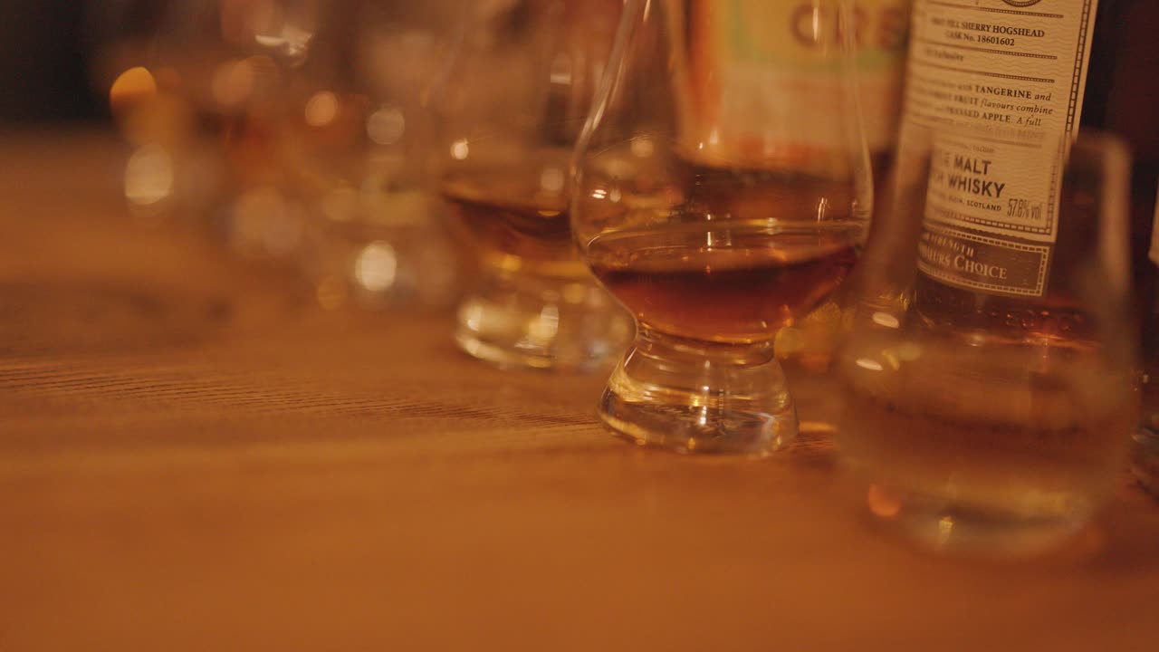 A hand reaches for a whisky glass among several tasting glasses on a warmly lit wooden table, suggesting a traditional whisky tasting setting