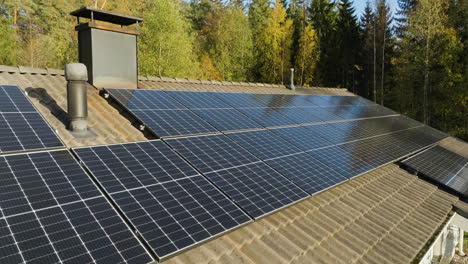 Aerial-view-of-roof-full-of-solar-panels-in-front-of-fall-foliage-trees,-sunny-day