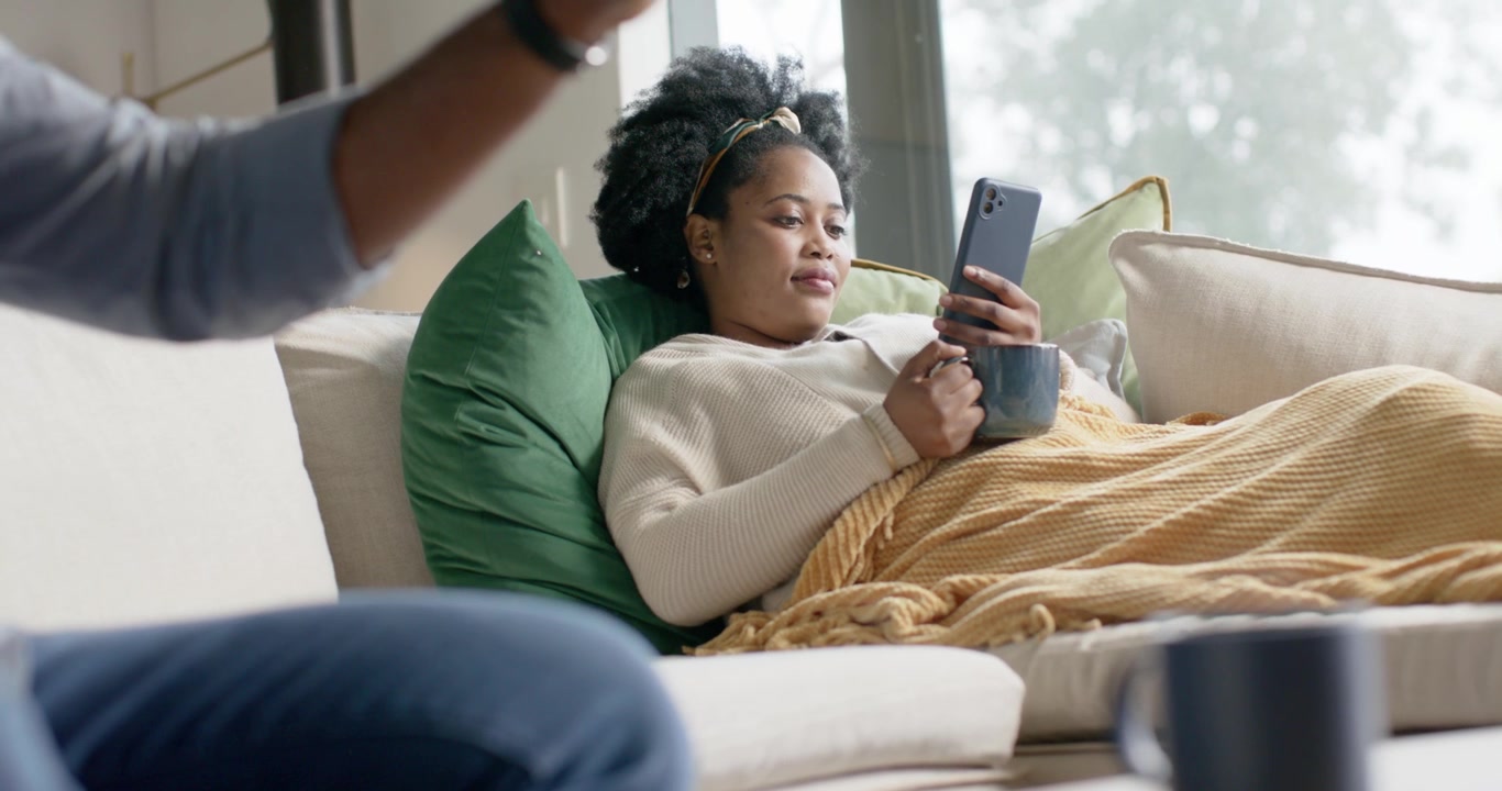 Donna afroamericana che si rilassa sul divano vicino alla finestra, usa il telefono e sorseggia da una tazza mentre delle mani maschili gesticolano.