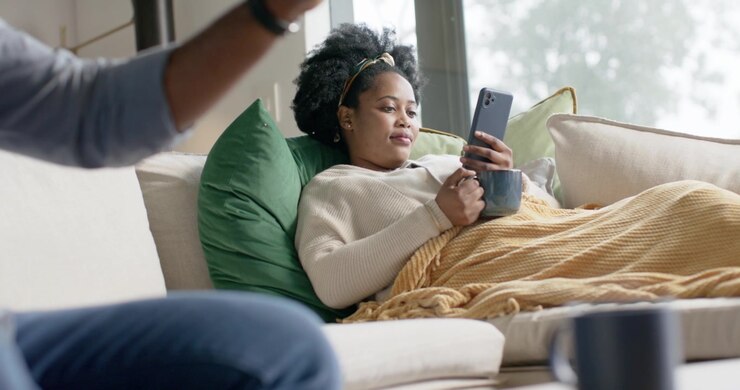 Donna afroamericana che si rilassa sul divano vicino alla finestra, usa il telefono e sorseggia da una tazza mentre delle mani maschili gesticolano.