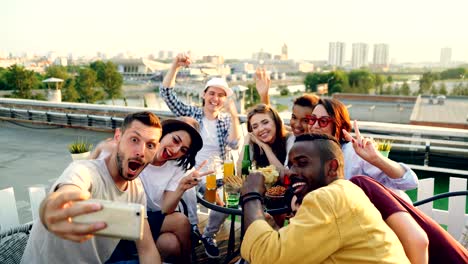 bearded guy is holding smartphone and taking selfie with friends who are posing with funny faces and hand gestures sitting at table on rooftop having fun.