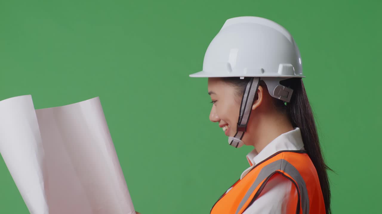 으로 가게 보이는 안전 헬을 입은 아시아 여성 엔지니어가 손에 청사진을 들고 녹색 스크린 배경 스튜디오에서 는 동안 주변을 둘러보고 있습니다.