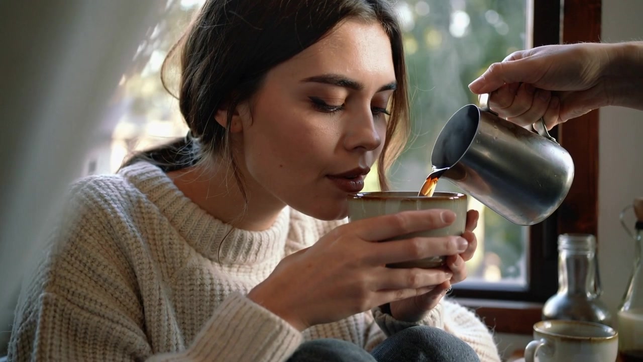 Donna con maglione di maglia che si appoggia e annusa il caffè al posto vicino al finestrino, mentre un'altra persona versa da una brocca in una tazza.