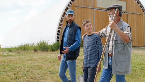 Side-view-of-grandfather,-father-and-grandson-farmers-walking-and-talking-in-green-field-while-the-sheep-flock-is-grazing