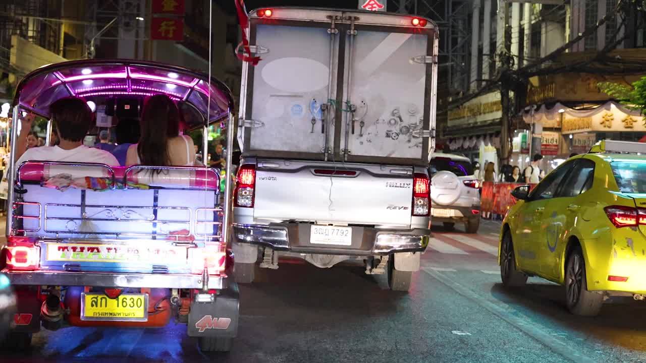 Couple travels by tuk-tuk under neon lights, passing busy traffic and vibrant street food stalls