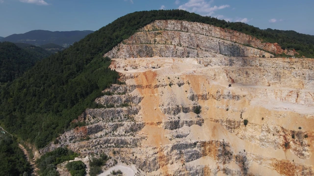 Ampio filmato aereo di una vasta cava a gradoni nella regione di Savinjska in Slovenia, con formazioni rocciose stratificate, foresta lussureggiante e luce diurna chiara.
