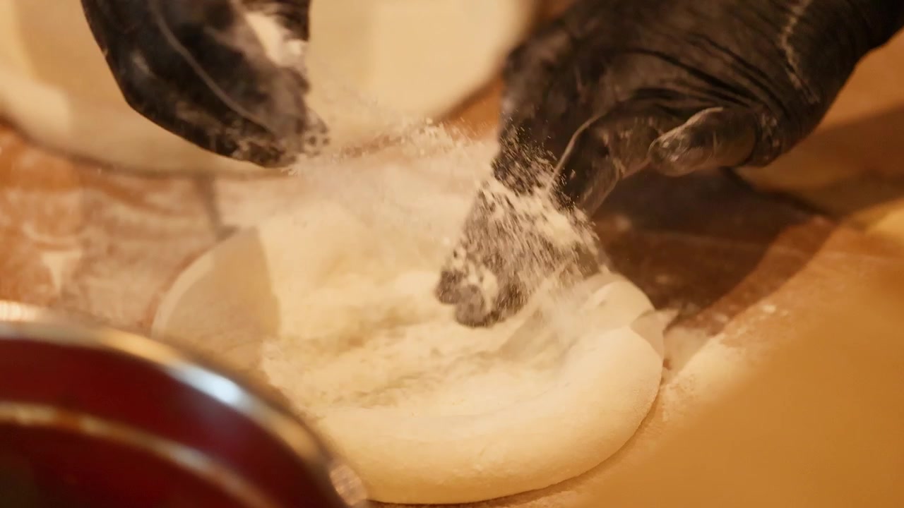 Close-up of hands in black gloves stretching fresh pizza dough on a floured surface.