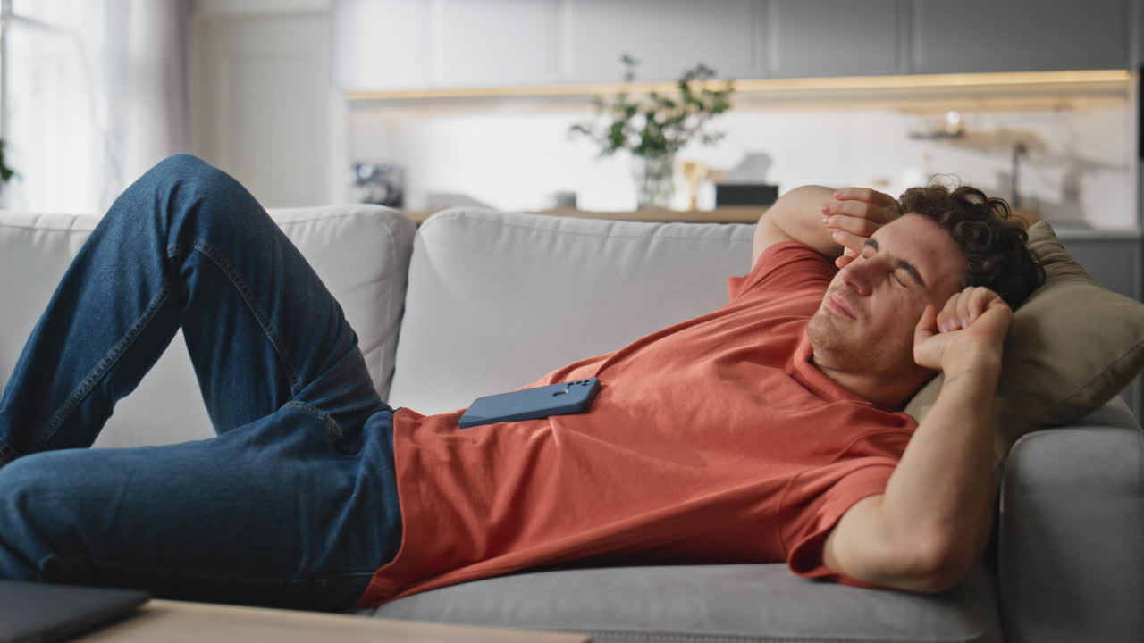 Lazy man browsing cellphone laying couch. Smiling guy putting down mobile phone