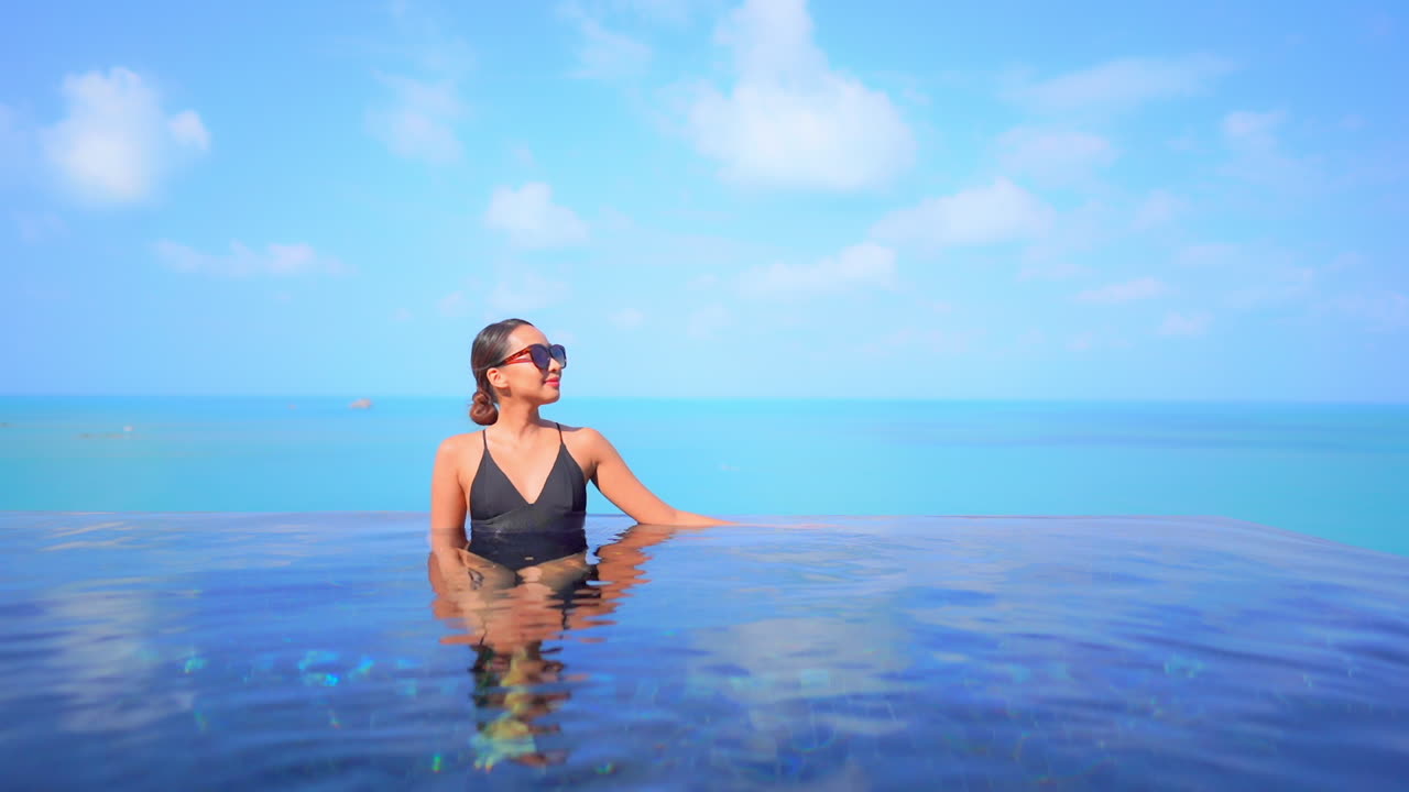 Fitte Thai-Frau im schwarzen Badeanzug entspannt sich am Rand eines Infinity-Pools vor türkisfarbener Meereskulisse, trägt Sonnenbrille an einem sonnigen Tag, Zeitlupe.