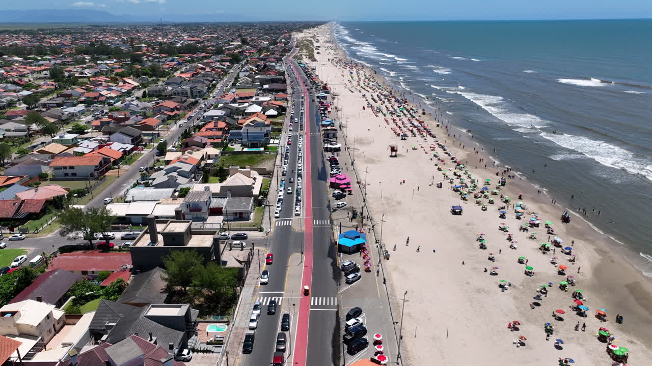 Vista aérea con dron de la concurrida playa de Imbé, con coloridas sombrillas, la costa con olas y el tráfico rodado con carril bici rojo en Río Grande del Sur, Brasil. Vídeo de vacaciones de verano en 4K a 60 FPS