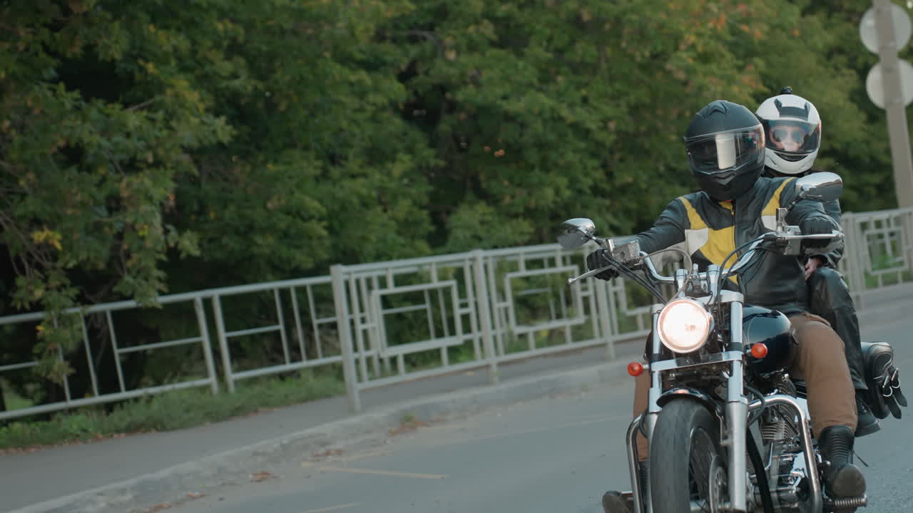 Motorcycle rider carrying passenger drives along tarred city road wearing helmets and leather jackets, chrome details reflecting light while passing green trees and roadside structure