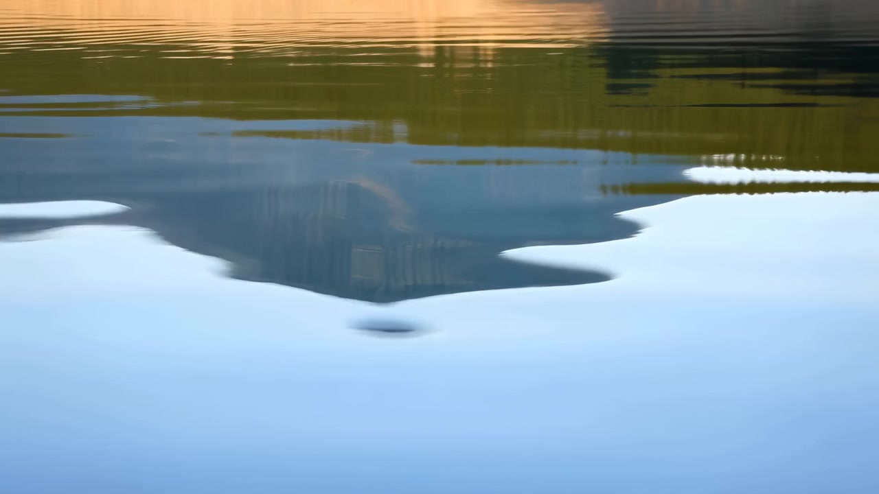 Panoramic close-up of lake surface with warm golden and olive reflections, darker mirrored landforms near center, pale blue water below, gentle ripples and soft shoreline detail, with reflection
