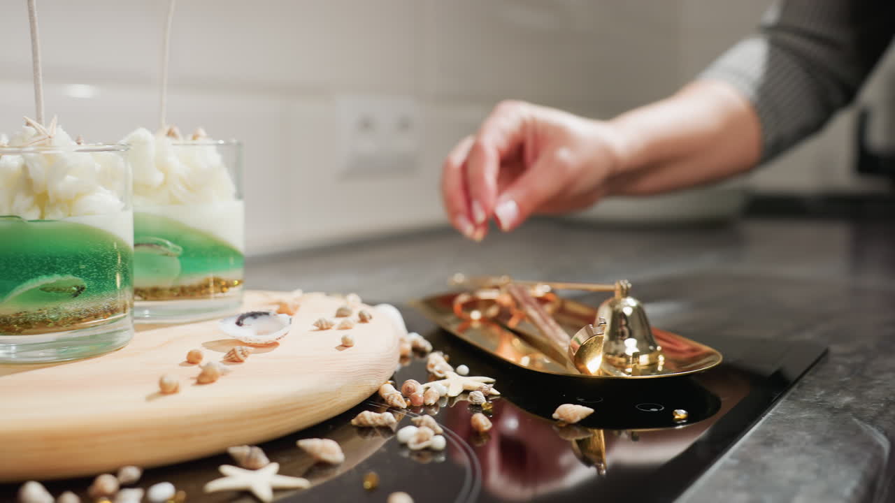 Female creative gently lifts golden tray containing scissors and candle tools, places it among elegant shell decorations on kitchen counter, preparing workspace for handcrafted candle styling