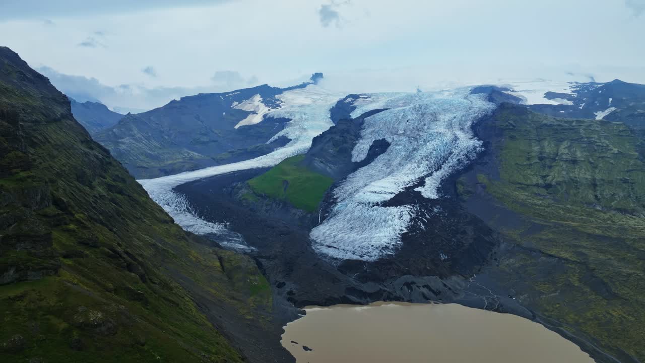 Dronepanorama af en islandsk udløbsgletsjer, der snor sig gennem en mørk vulkansk dal mod en gulbrun smeltevandslagune, indrammet af grønne skråninger og forrevne bjerge.